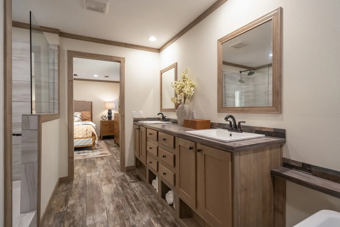 Bathroom with dual sinks, wood cabinetry, large mirrors, and a view into a bedroom with a bed and nightstand in the background.
