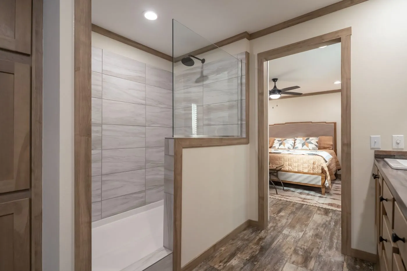 Modern bathroom with a walk-in shower featuring a partial glass wall and wood trim, adjacent to a bedroom with a ceiling fan and patterned bedding.