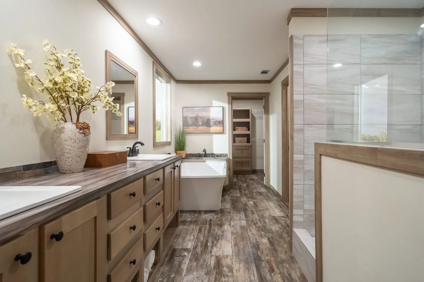 Modern bathroom with wood cabinets, double sinks, a freestanding tub, decorative vase, and a glass-enclosed shower, featuring wood-look flooring and neutral tones.