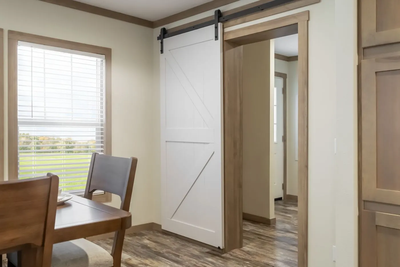 Dining area with wooden chairs and table next to a large window; a white barn-style sliding door is partially open, revealing a hallway with wood flooring.