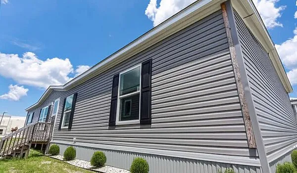 Gray single-story manufactured home with vinyl siding, black shutters, and a small front porch, set on a grassy lot with trimmed shrubs. Blue sky with scattered clouds above.